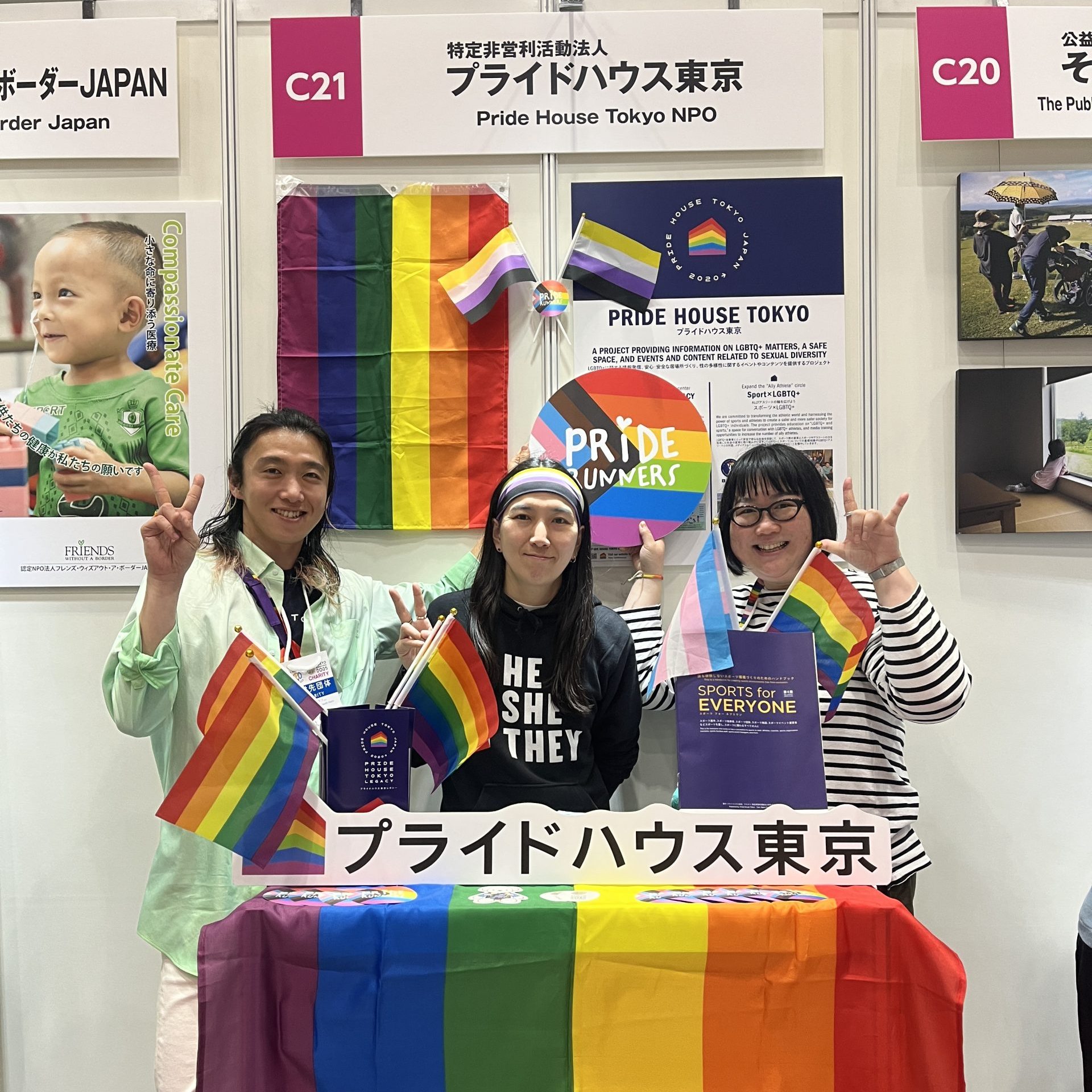 プライドハウス東京の東京マラソンEXPO 2025チャリティブースの写真。 スタッフが三人並んでいる。 ブースはプライドフラッグやノンバイナリー・プライド・フラッグで飾られている。